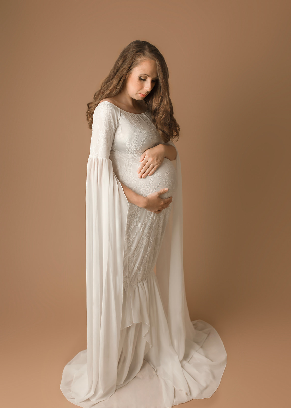 A mother to be stands in a studio looking down at her bump in her hands in a white maternity gown before a 3D Ultrasound in Houston