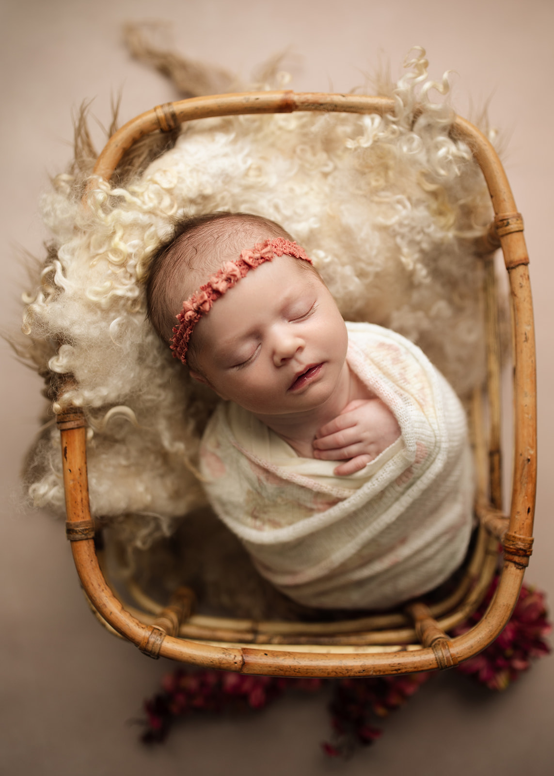 A newborn baby sleeps in a wicker bucket in a white swaddle and pink headband