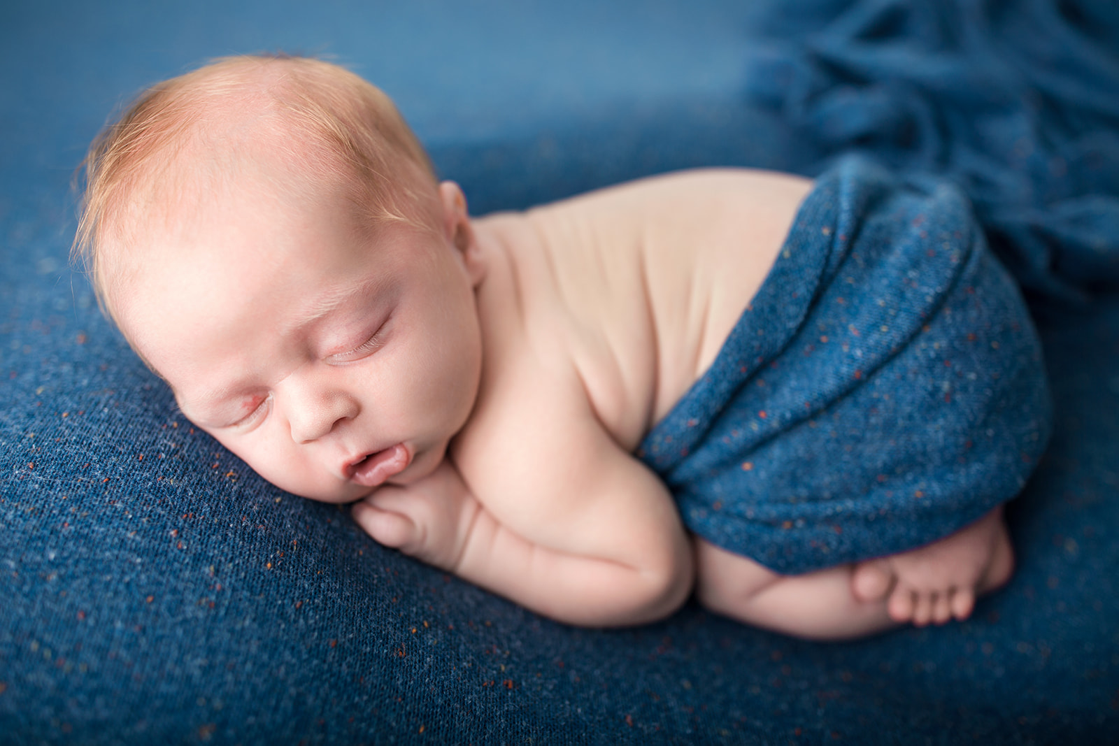 A newborn baby sleeps wrapped in a blue blanket on a matching bed in a studio