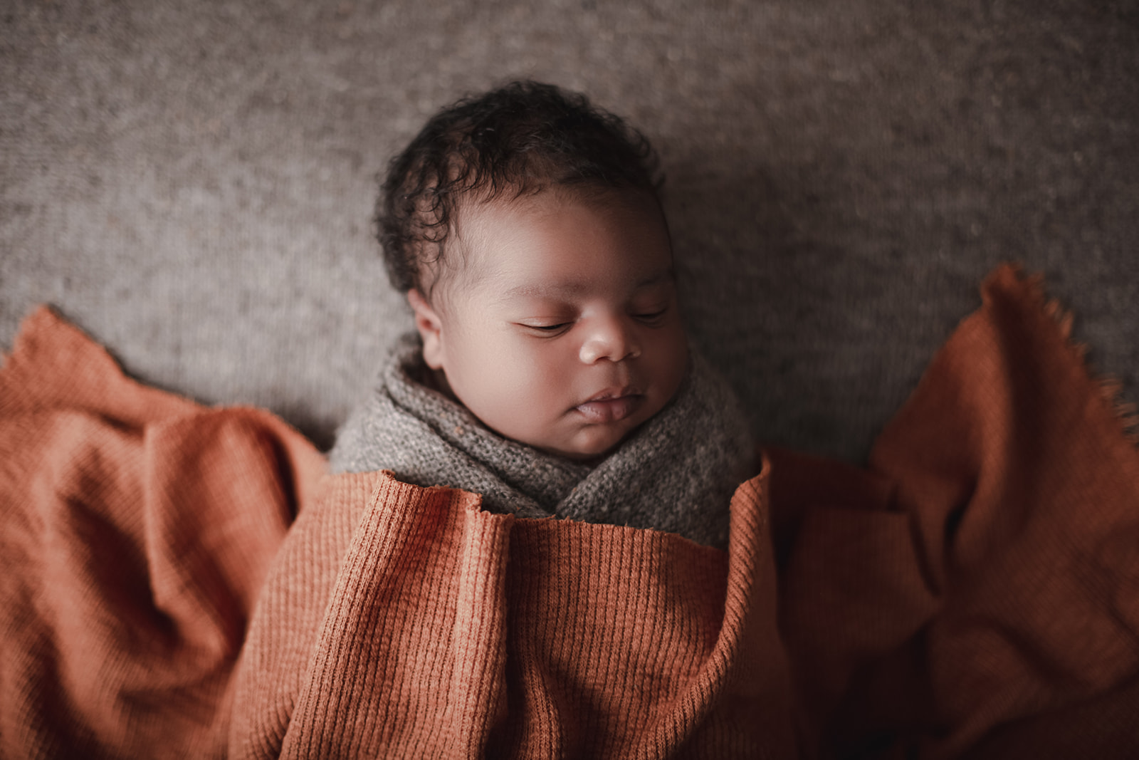 A newborn baby sleeps in a grey knit swaddle under an orange blanket