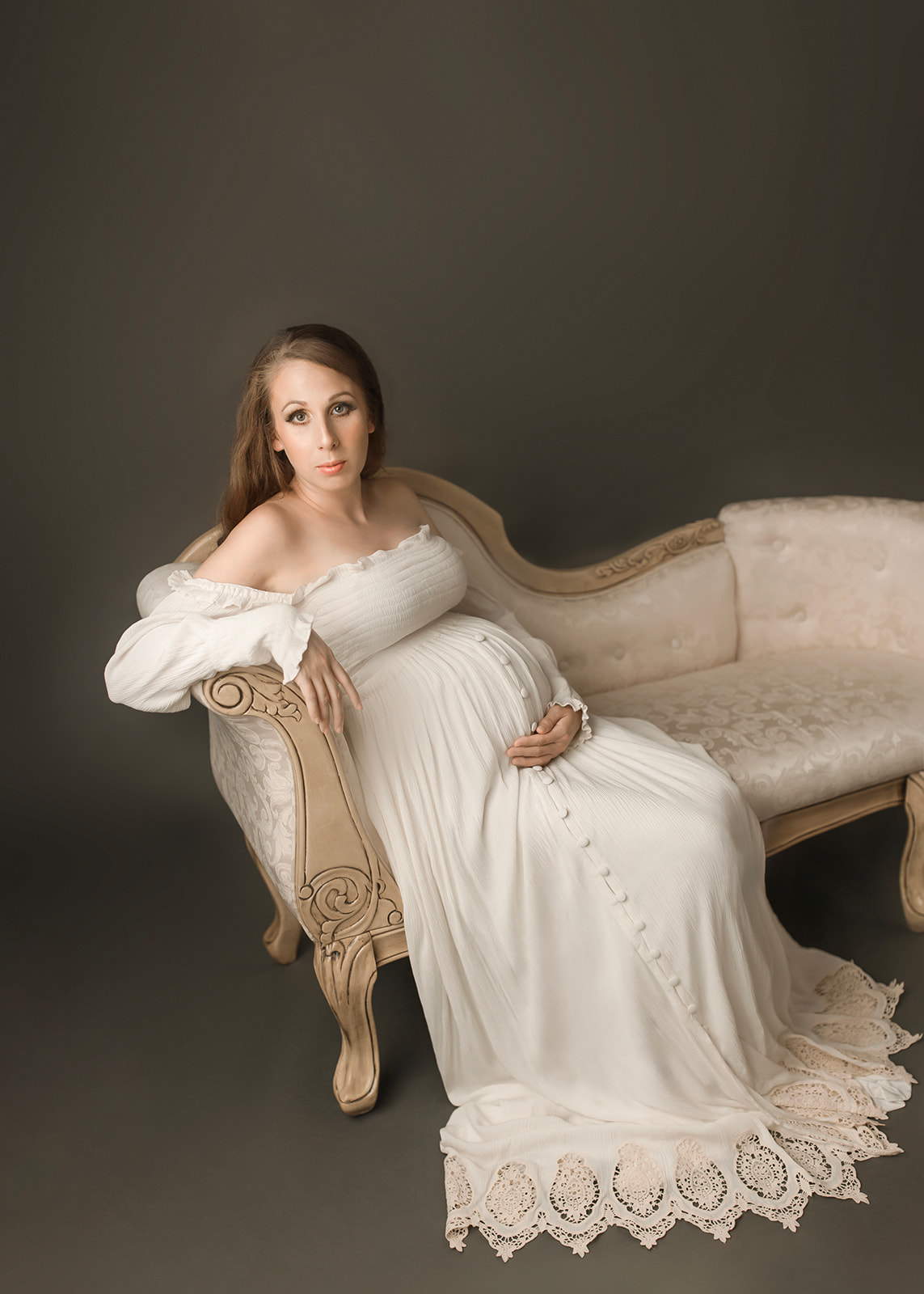 A mom to be sits on an antique couch with a hand under her bump in a white maternity gown