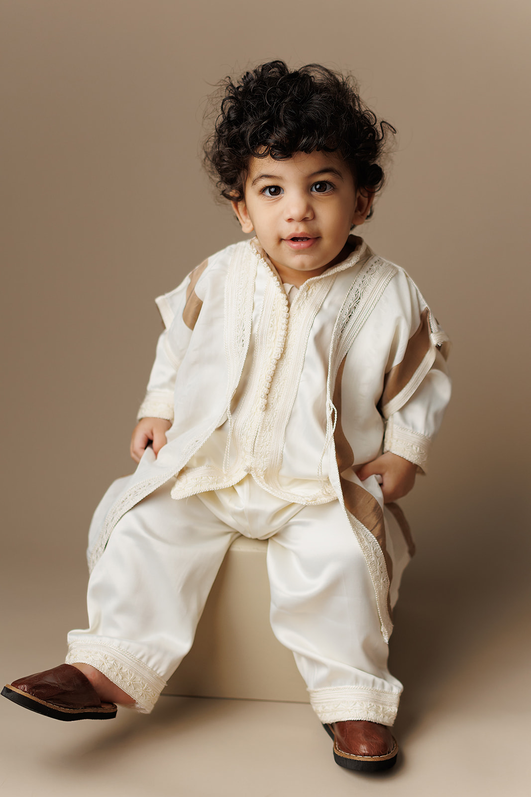 A young toddler in a traditional white outfit sits on a box in a studio after visiting Houston Daycares