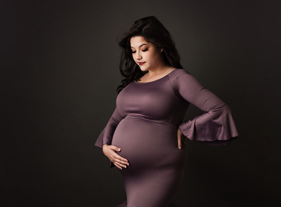 A mother to be stands in a studio in a dark purple maternity gown smiling down to her bump