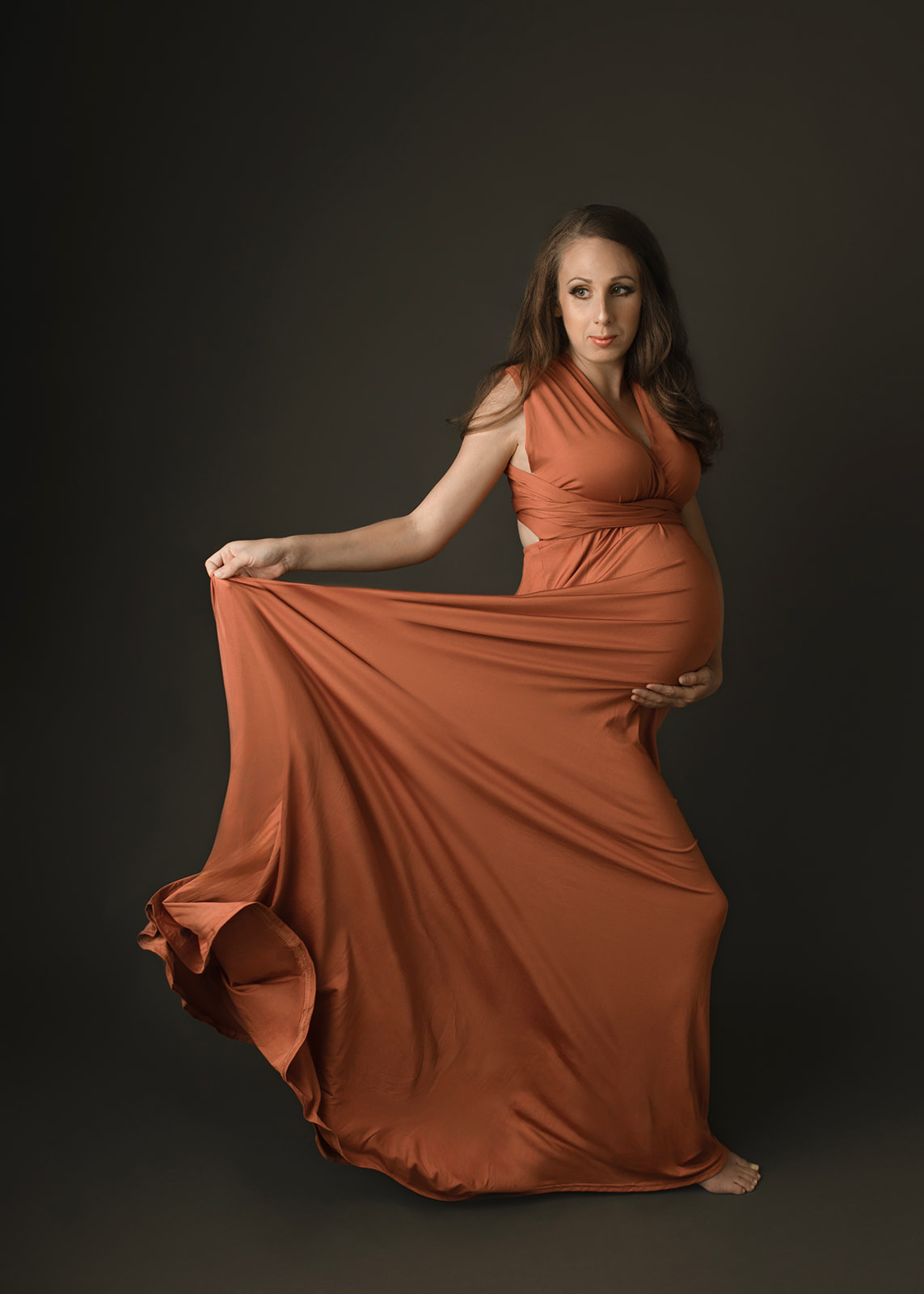 A mother to be in an orange maternity gown stands in a studio playing with her train and holding the bump after meeting Houston Doulas