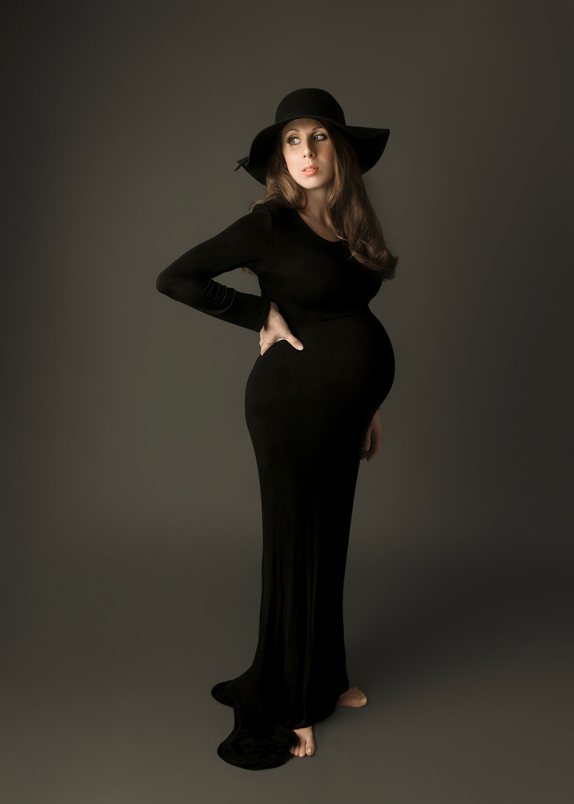 A mother to be stands barefoot in a sstudio in a black maternity gown and matching hat looking over her shoulder after meeting Houston Doulas