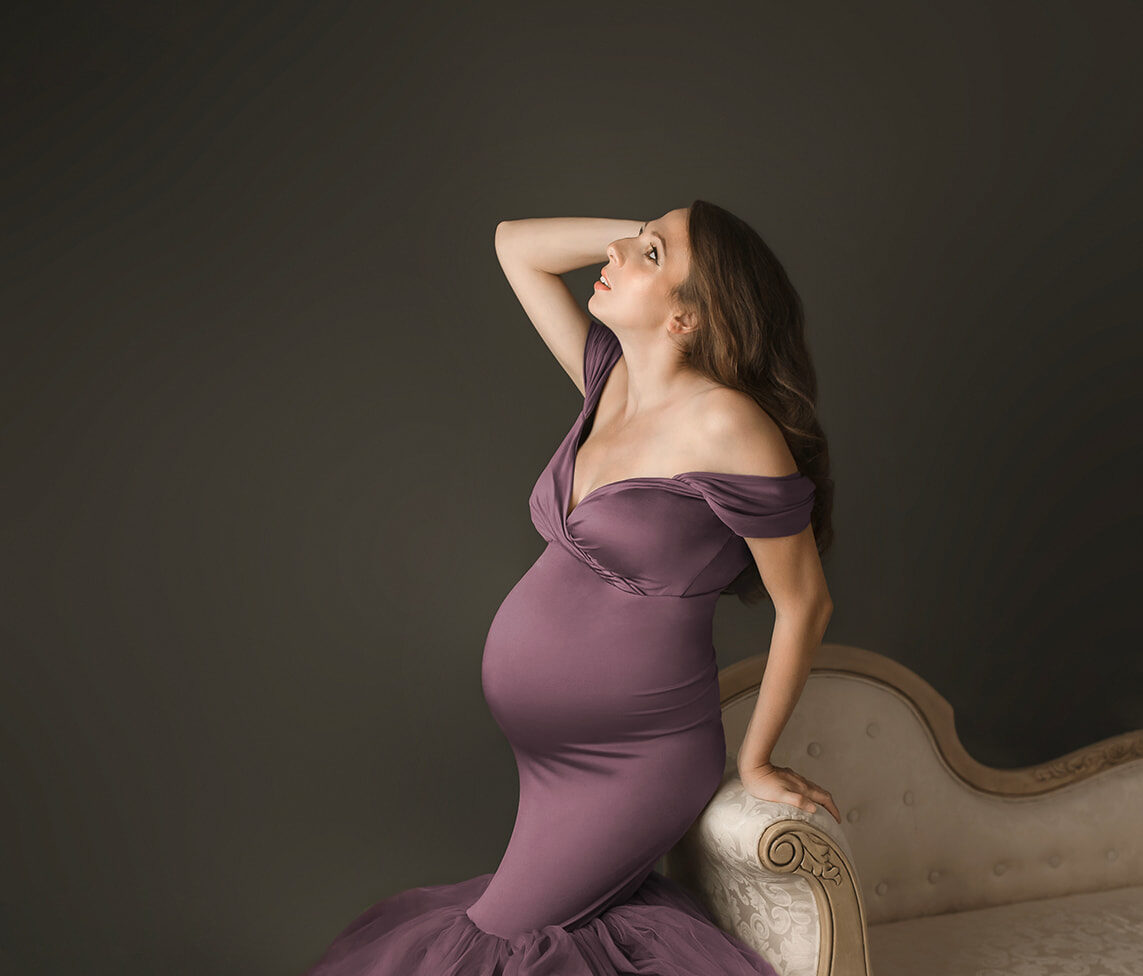 A mother to be in a purple maternity gown leans on a vintage couch while looking up with a hand in her hair after meeting Houston Midwives