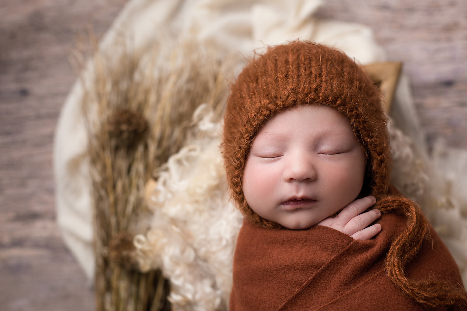 A newborn baby sleeps in a brown knit bonnet and matching swaddle in a studio