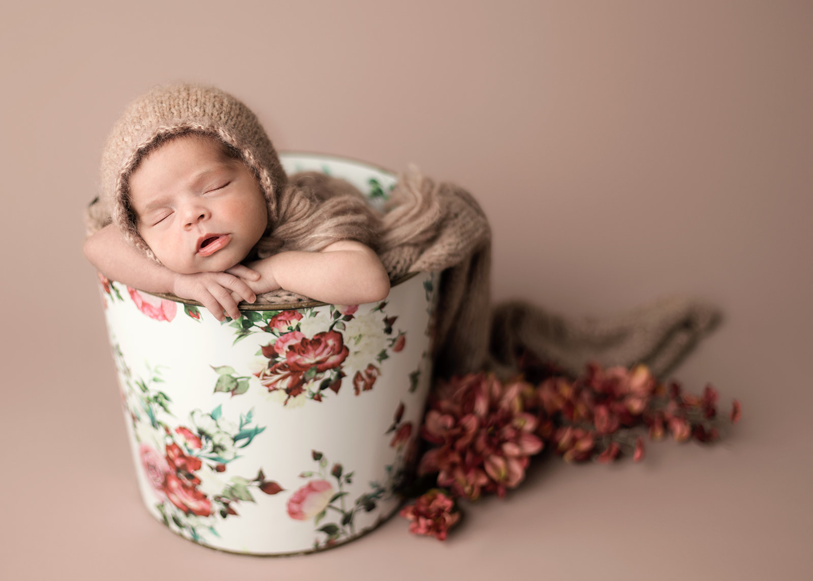 A newborn baby sleeps in a metal rose printed bucket in a tan knit bonnet in a studio thanks to Houston Postpartum Doulas