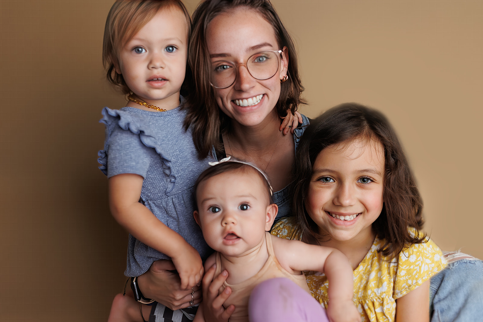 A happy mother sits on the floor of a studio with her three toddler daughters in her lap hugging her