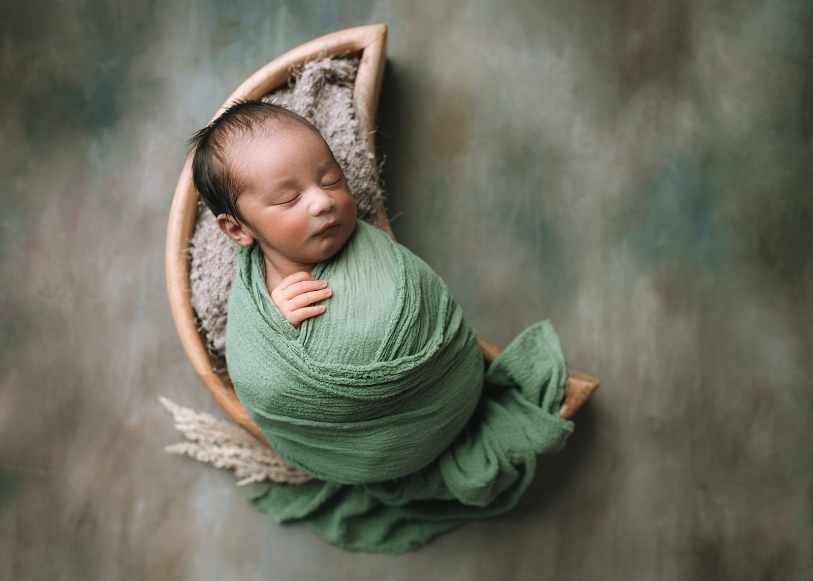 A newborn baby in a green swaddle sleeps in a moon shaped wooden bowl