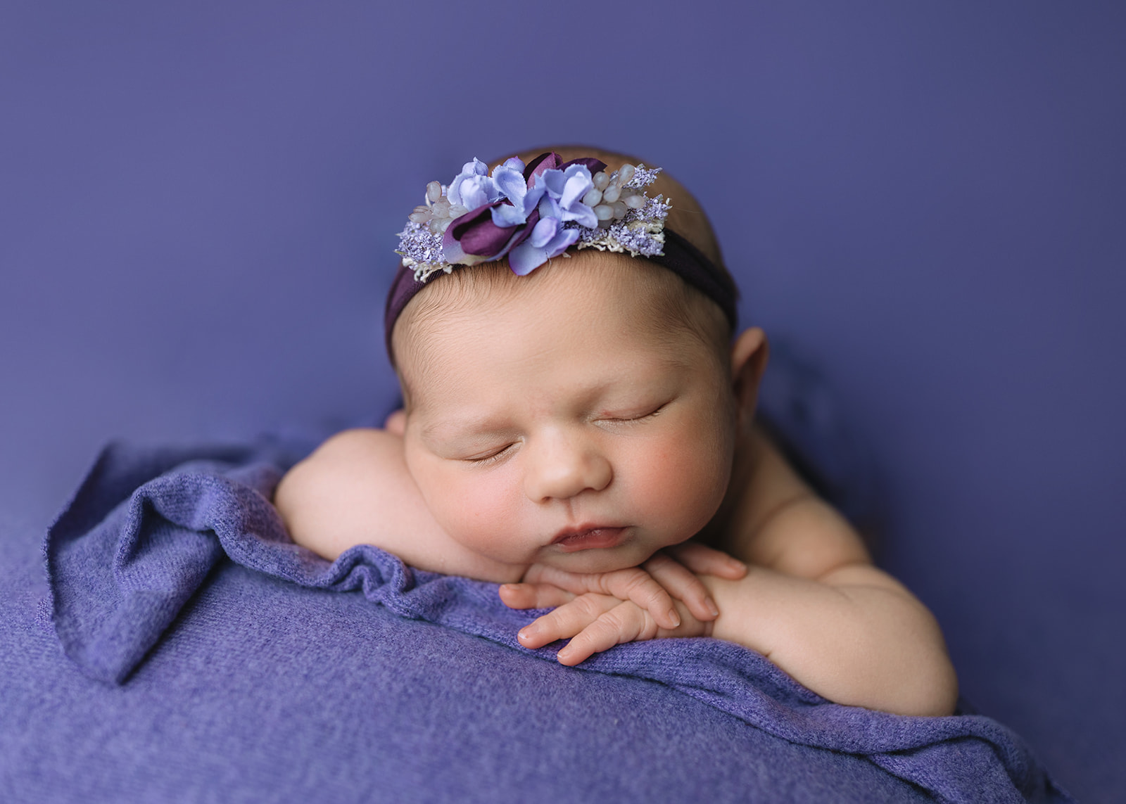 A newborn baby girl in a purple floral headband sleeps on a purple blanket in a studio after visiting Houston Baby Boutiques