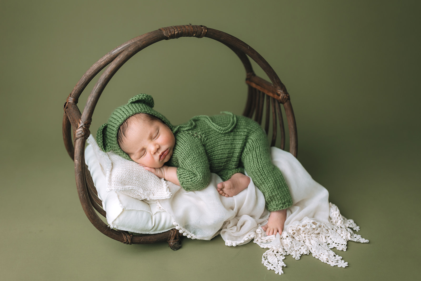 A newborn baby sleeps on a circular wooden wicker bed in a green knit onesie from Houston Baby Boutiques