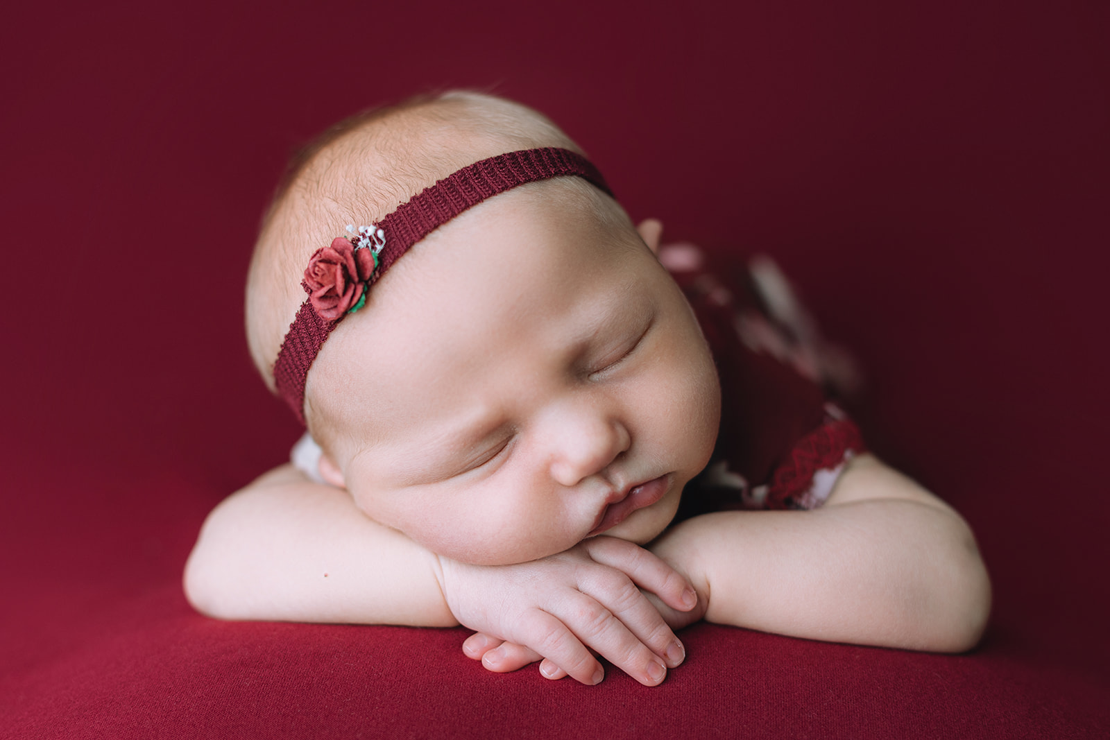 A newborn baby in a red rose headband sleeps on her hands in a studio