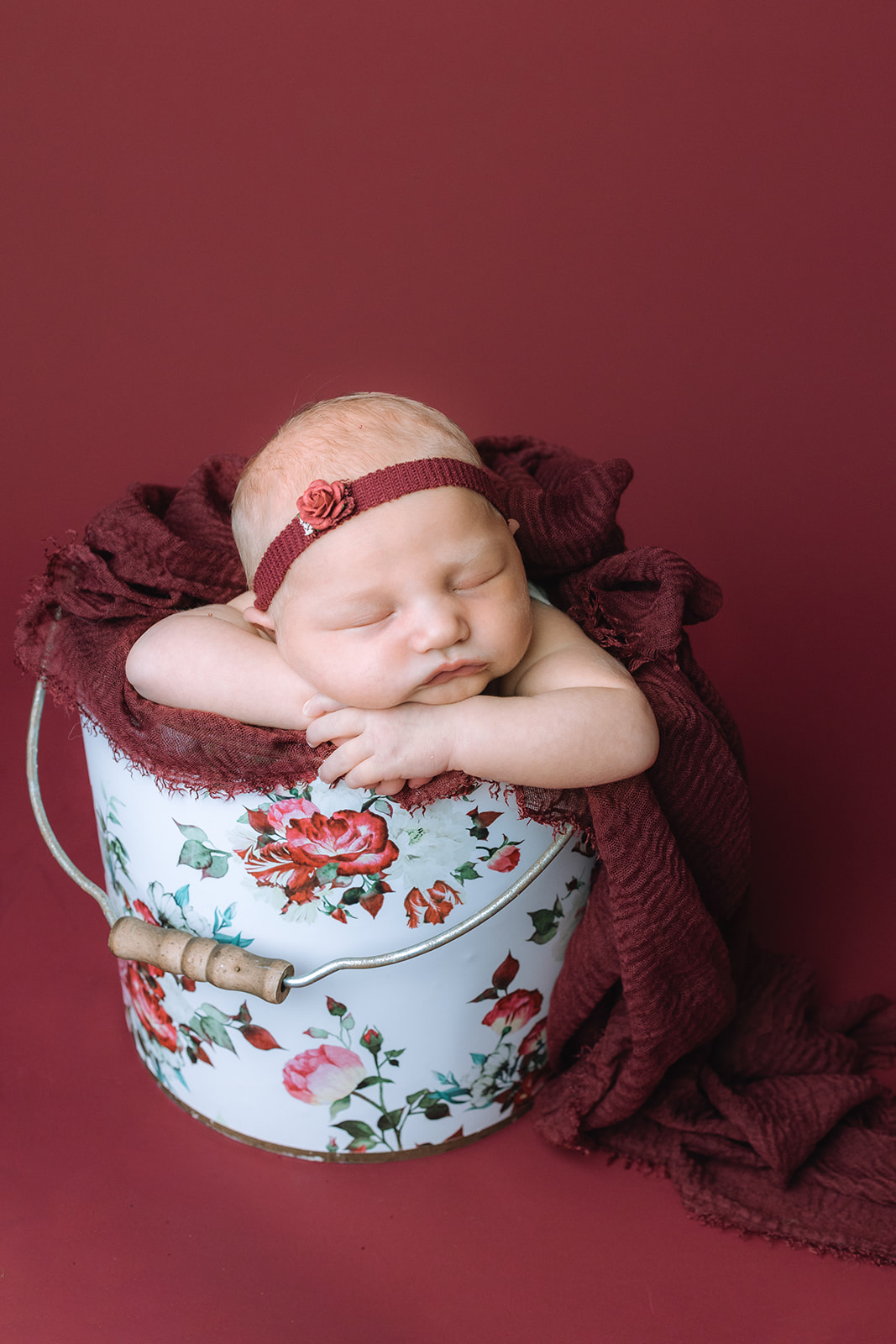 A newborn baby girl in a red headband sleeps in a floral print bucket in a studio thanks to Houston lactation consultants