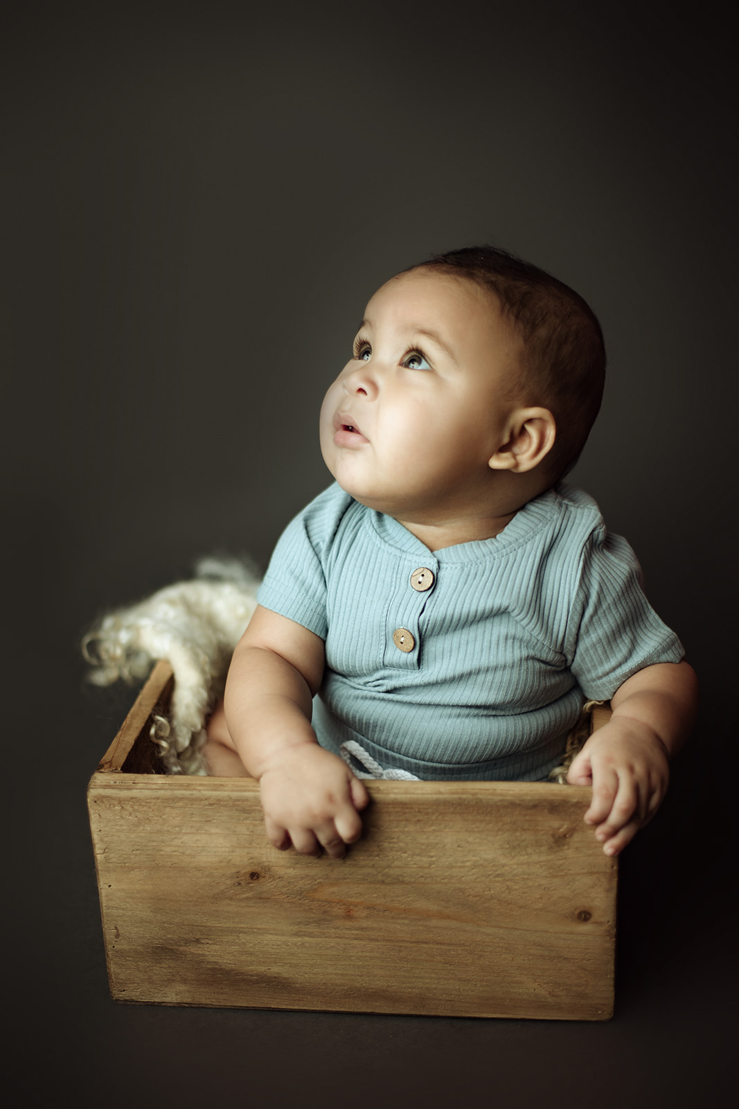 An infant boy in a blue onesie sits in a wooden box in a studio