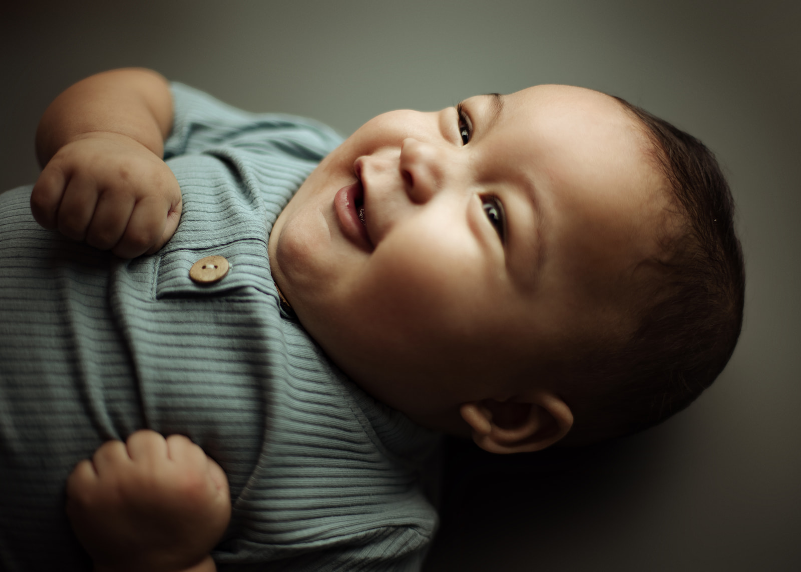 An infant boy in a blue onesie laughs while laying on a bed in a studio after meeting Houston Pediatric Dentists