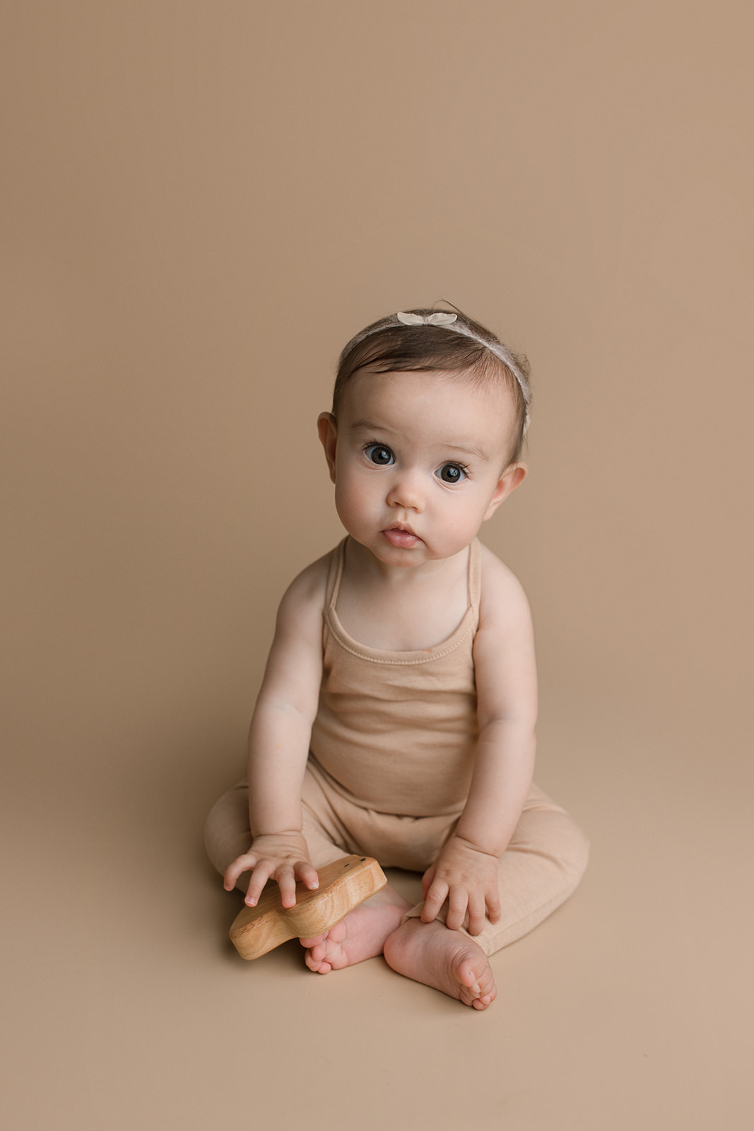 A toddler girl sits on the floor of a studio playing with a wooden toy in a brown onesie