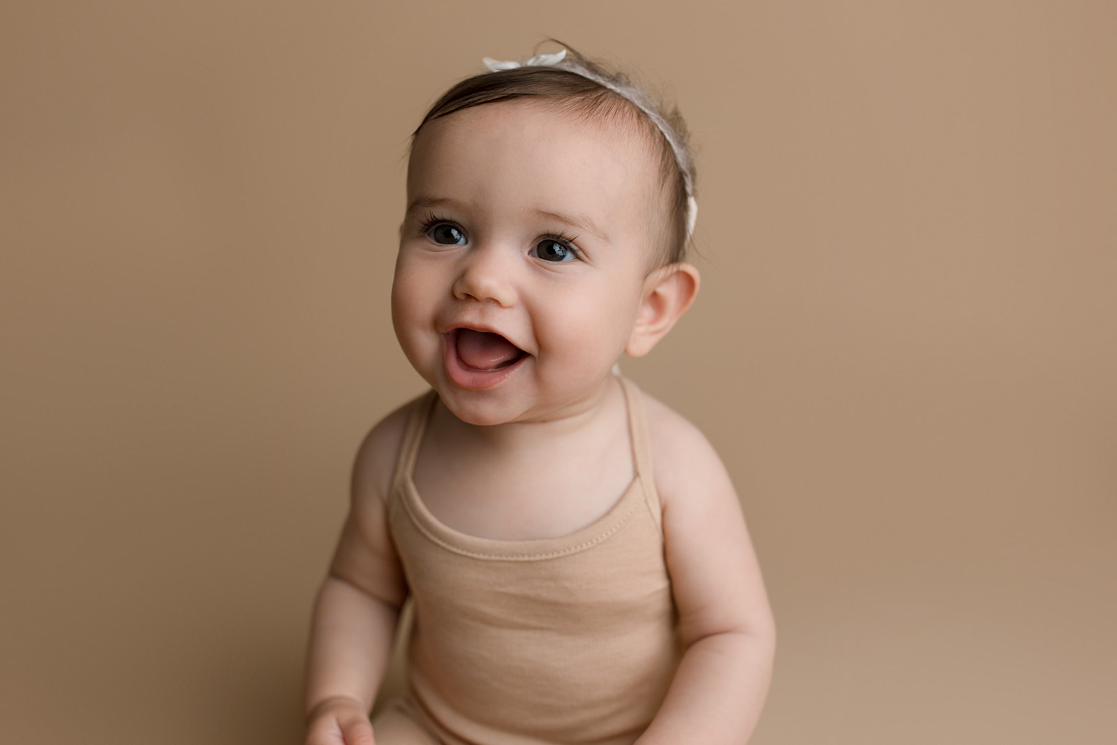 A young toddler girl smiles big while sitting in a studio in a brown onesie and white bow after visiting Houston Pediatricians