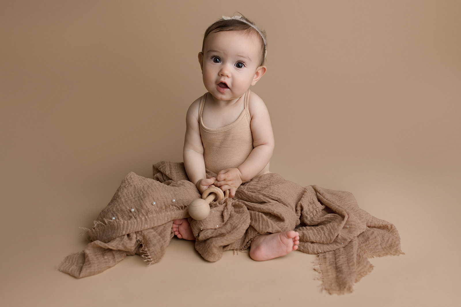 A toddler girl sits in a brown blanket playing with a wooden toy in a studio after meeting Houston Pediatricians