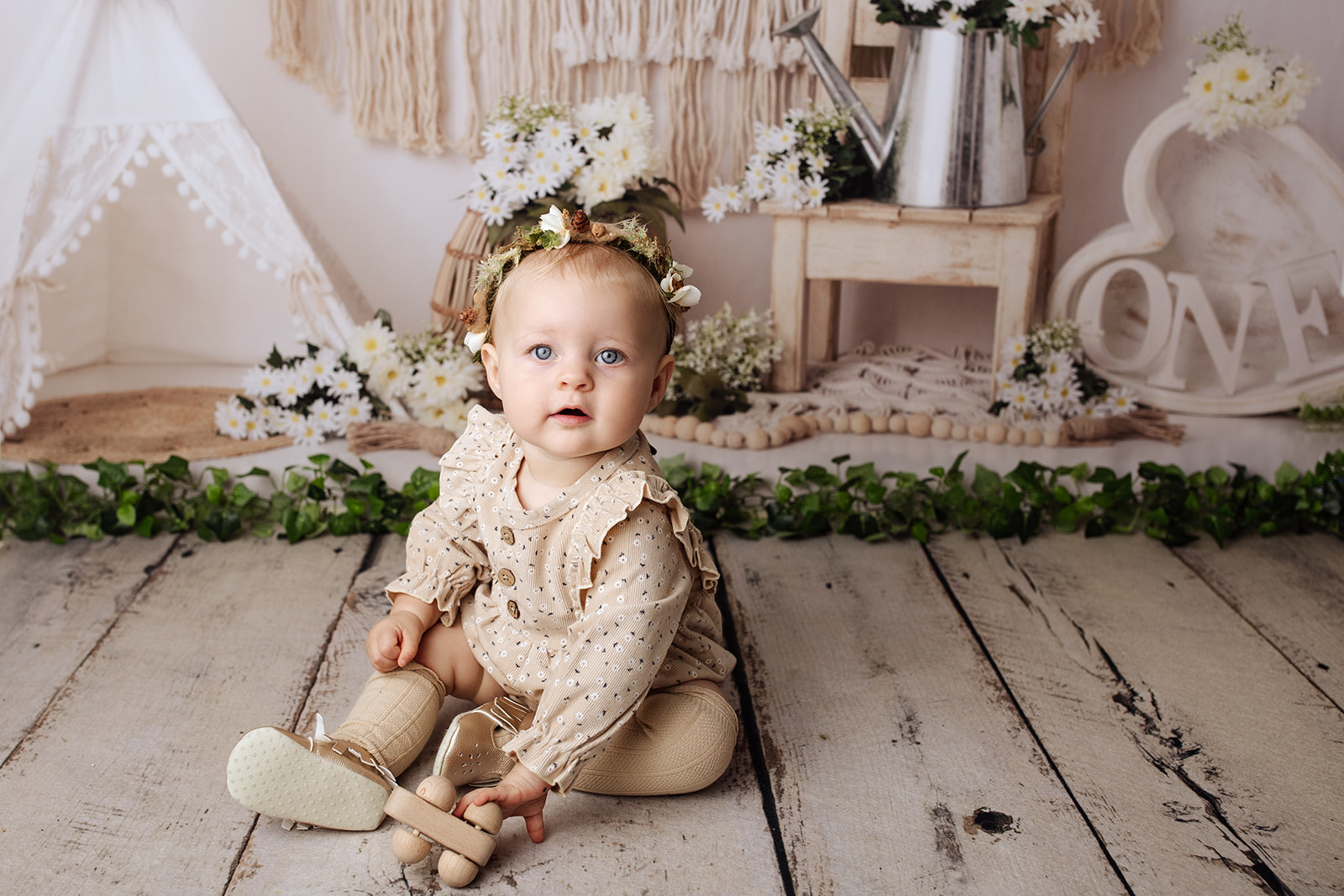 A young toddler girl in a brown print dress onesie plays with a wooden toy on the floor of a studio for her first birthday