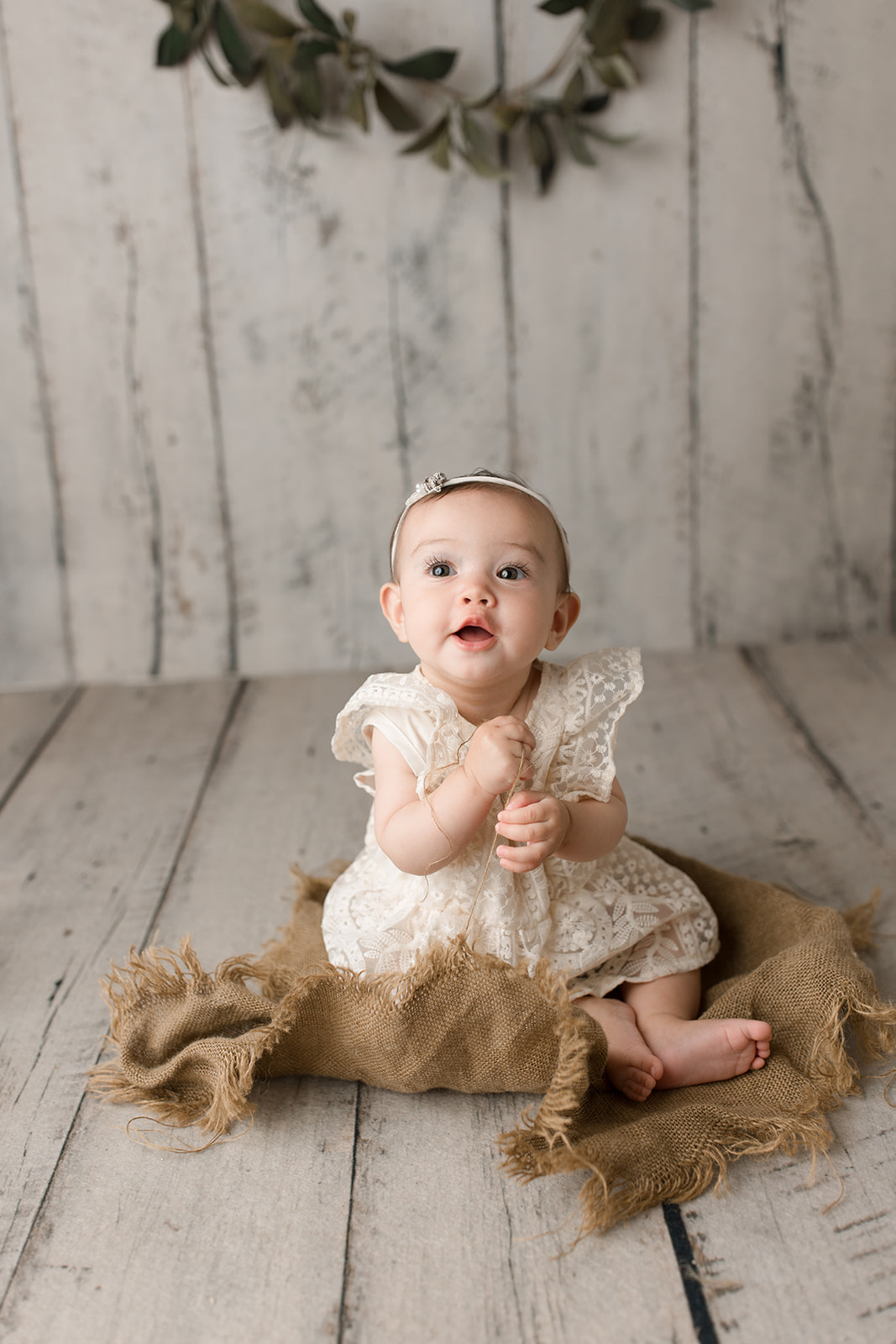 An infant girl in a white lace dress sits on the floor of a studio on a burlap blanket thanks to Houston Prenatal Massage