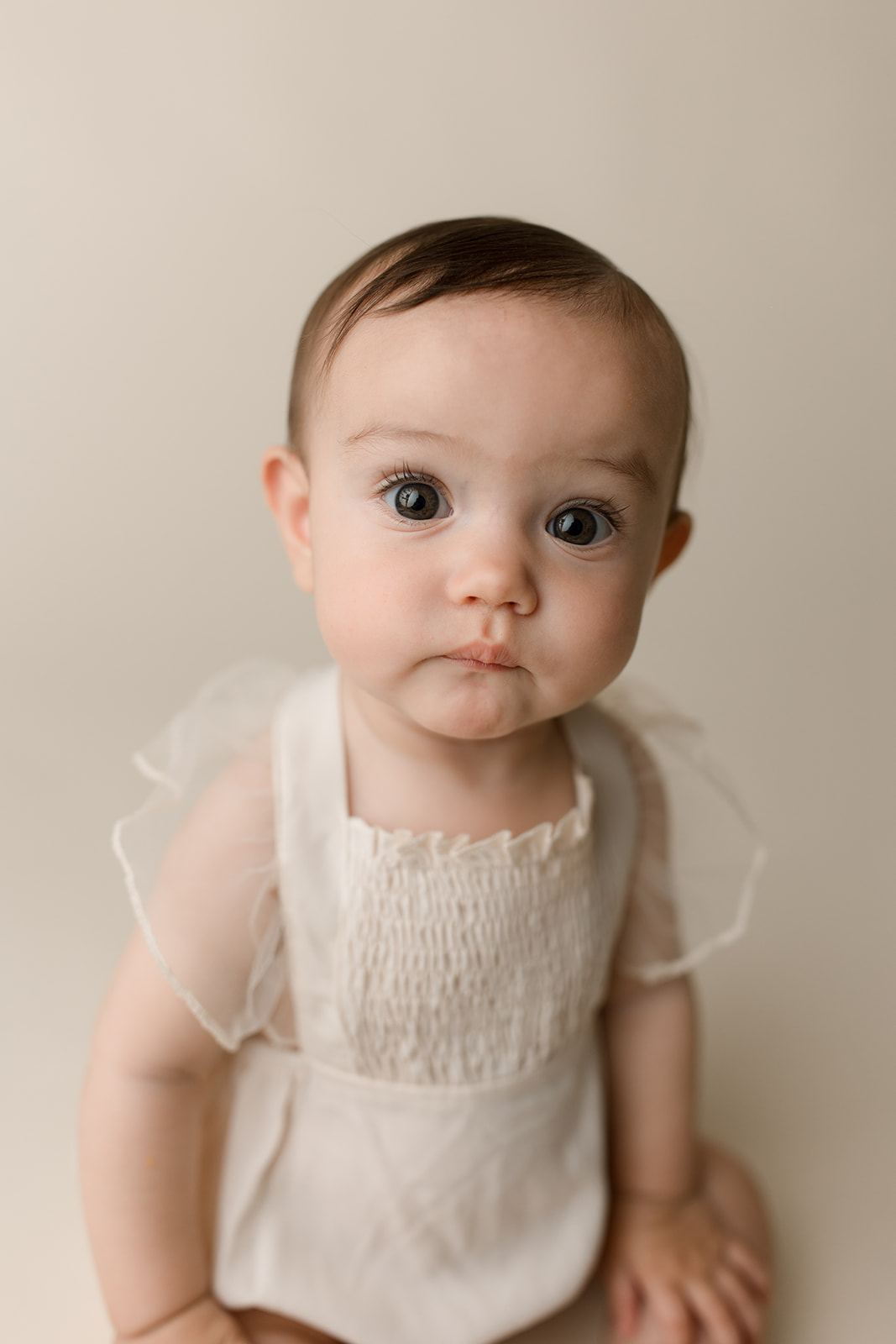 An infant girl sits on the floor of a studio in a white dress with a curious face after mom used Houston Prenatal Massage