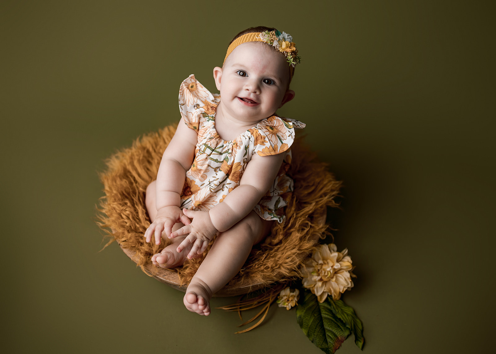 An infant girl in an orange floral print dress sits in a basket with a matching blanket in a studio