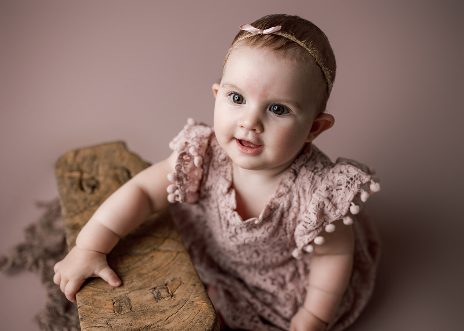 A young infant girl in a pink dress plays with a wooden stool in a studio