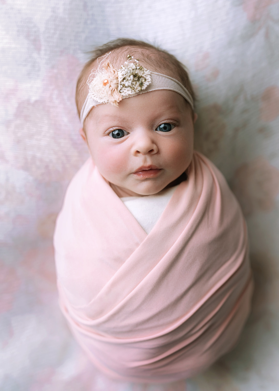 A newborn baby lays on a bed in a pink swaddle and floral headband with eyes open thanks to OBGYN In Houston