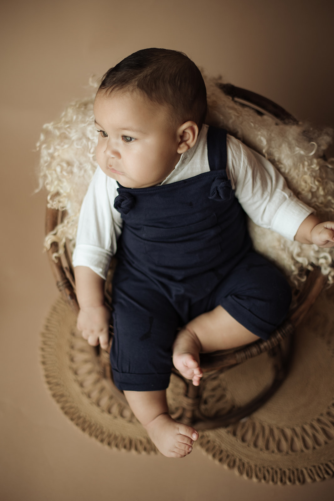 A young infant sits in a wicker chair in a studio in blue overalls after some Parenting Classes Houston