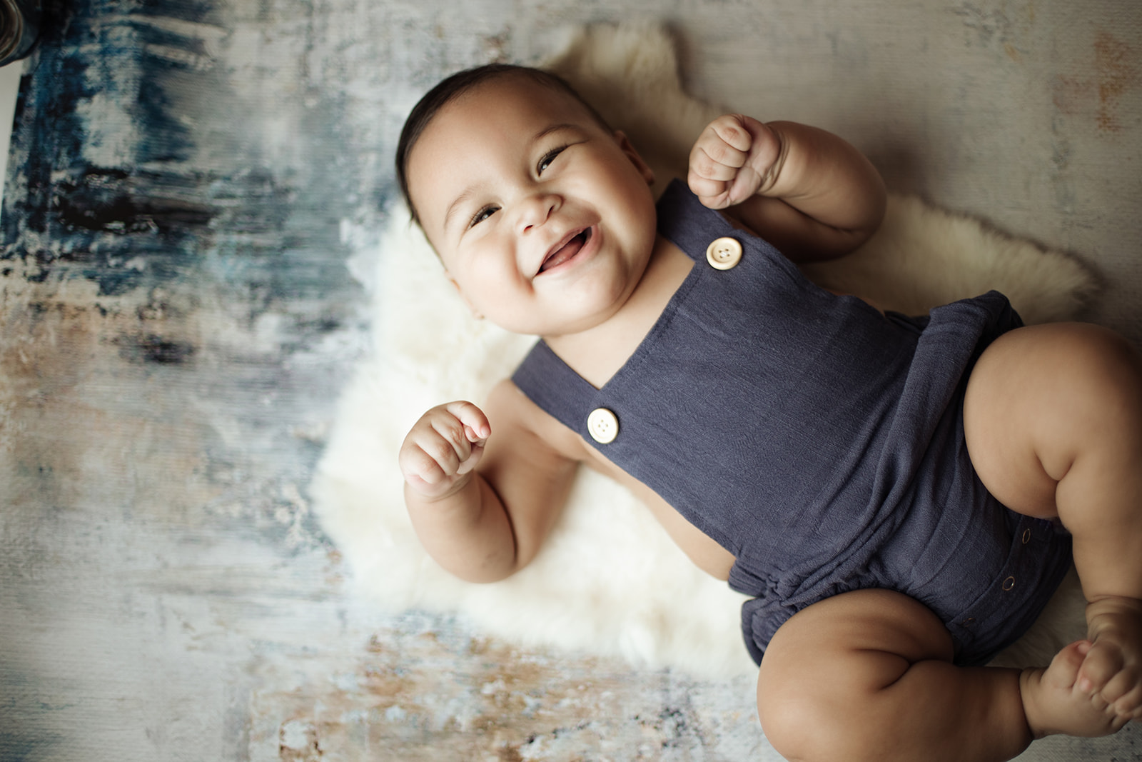 An infant lays on a rug smiling big in blue overalls