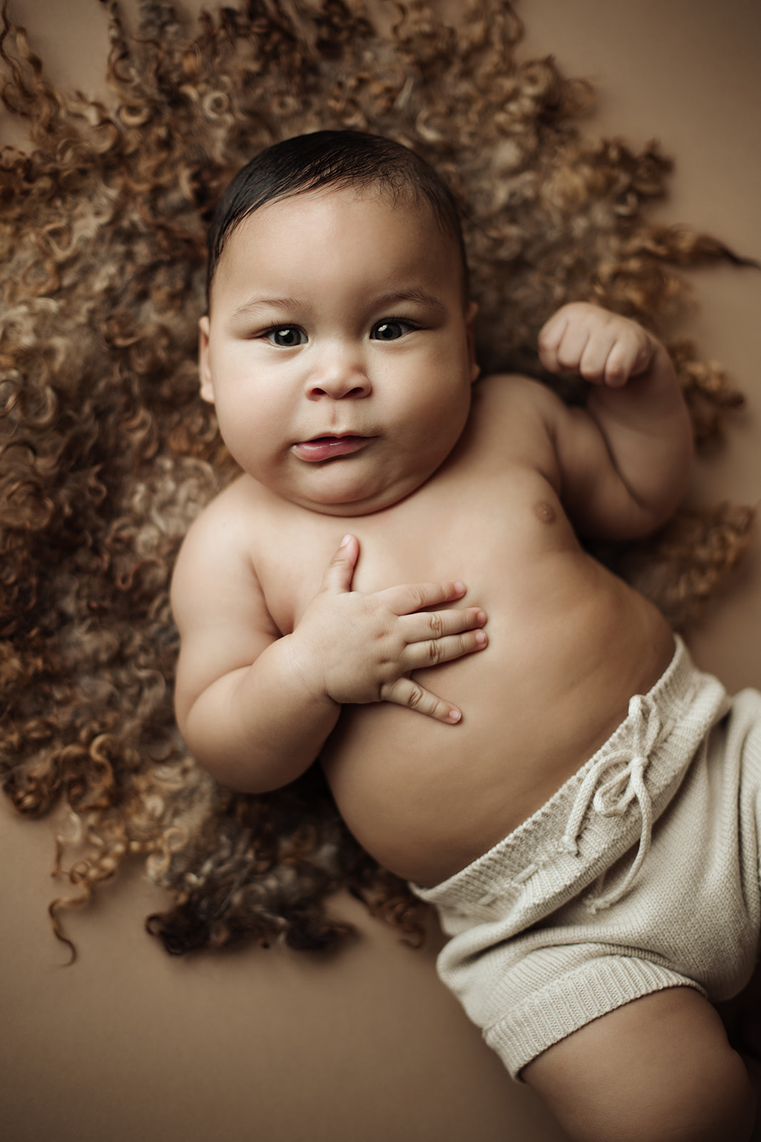 A newborn baby in tan shorts lays on a brown blanket with a hand on it's belly thanks to a Prenatal Chiropractor Houston