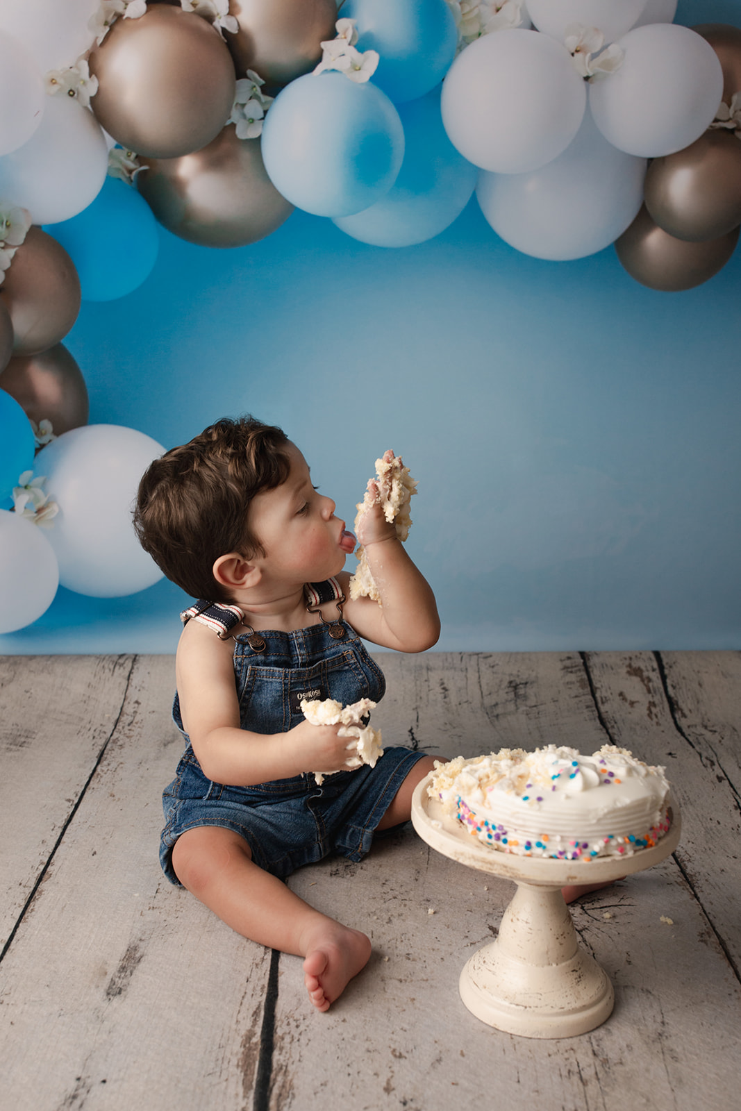 A toddler boy in denim overalls licks cake off his hands in a studio for his cake smash session after some baby music classes in Houston