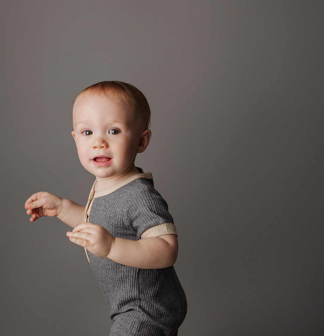 A young toddler stands up from a wooden stool in a grey onesie in a studio after using a Houston diaper service