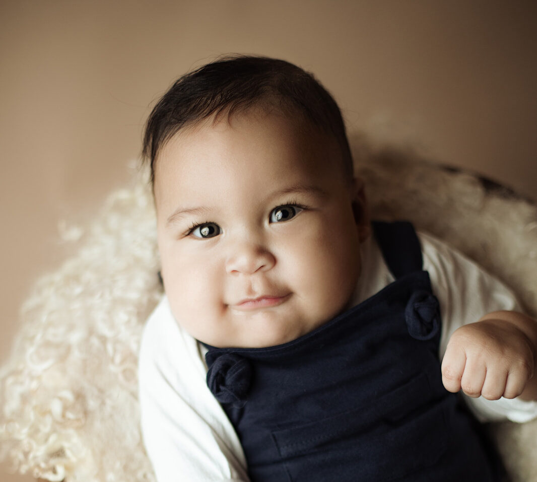 An infant boy lays in a wicker chair in blue overalls playing with a wooden toy while mom gets Pelvic Floor Therapy Houston