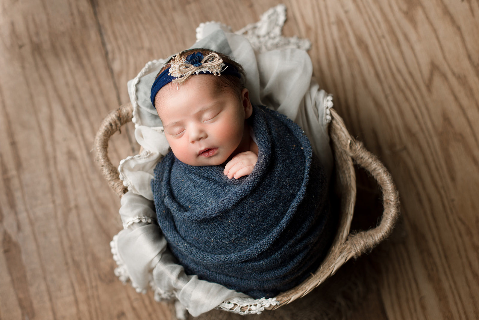 A newborn baby girl sleeps in a woven basket while wrapped in a navy blue knit swaddle