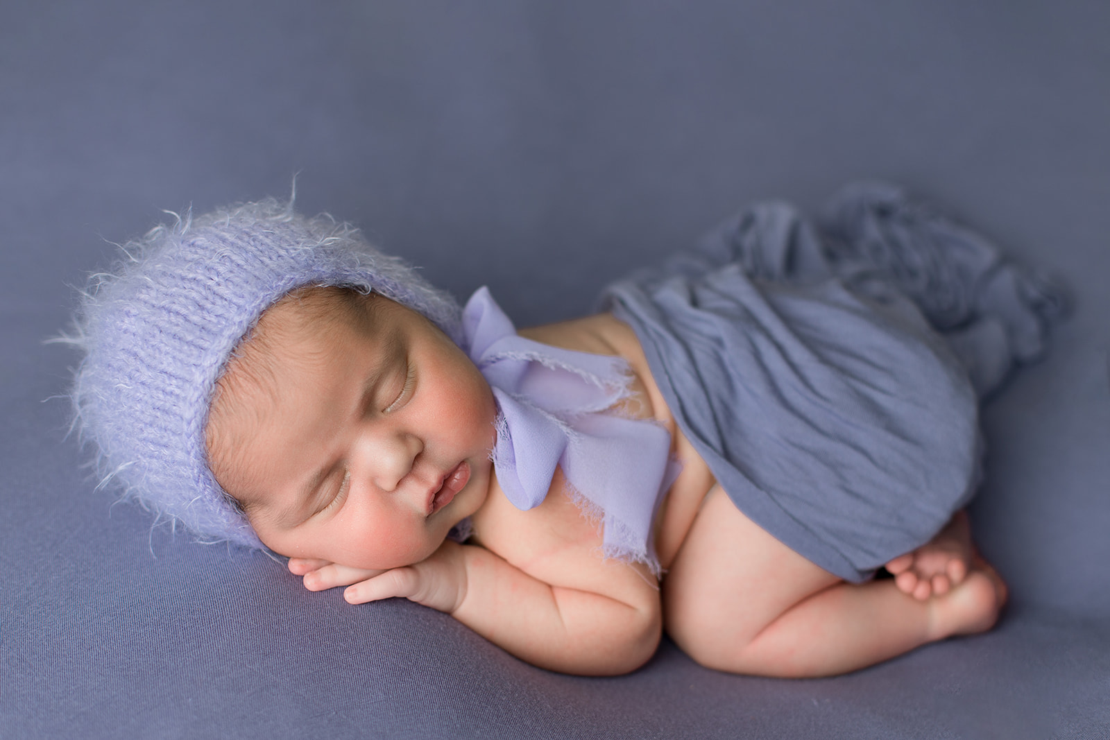 A newborn baby in a blue knit bonnet sleeps on its stomach in a studio