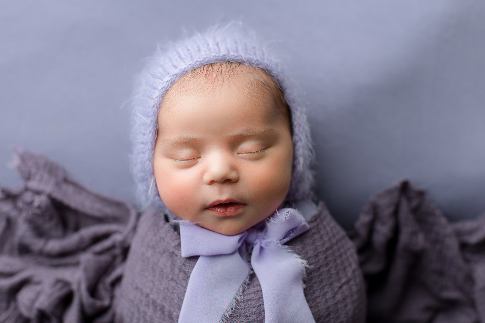 A sleeping newborn baby lays wrapped in a tight purple swaddle with a blue knit bonnet after meeting Houston Babysitters