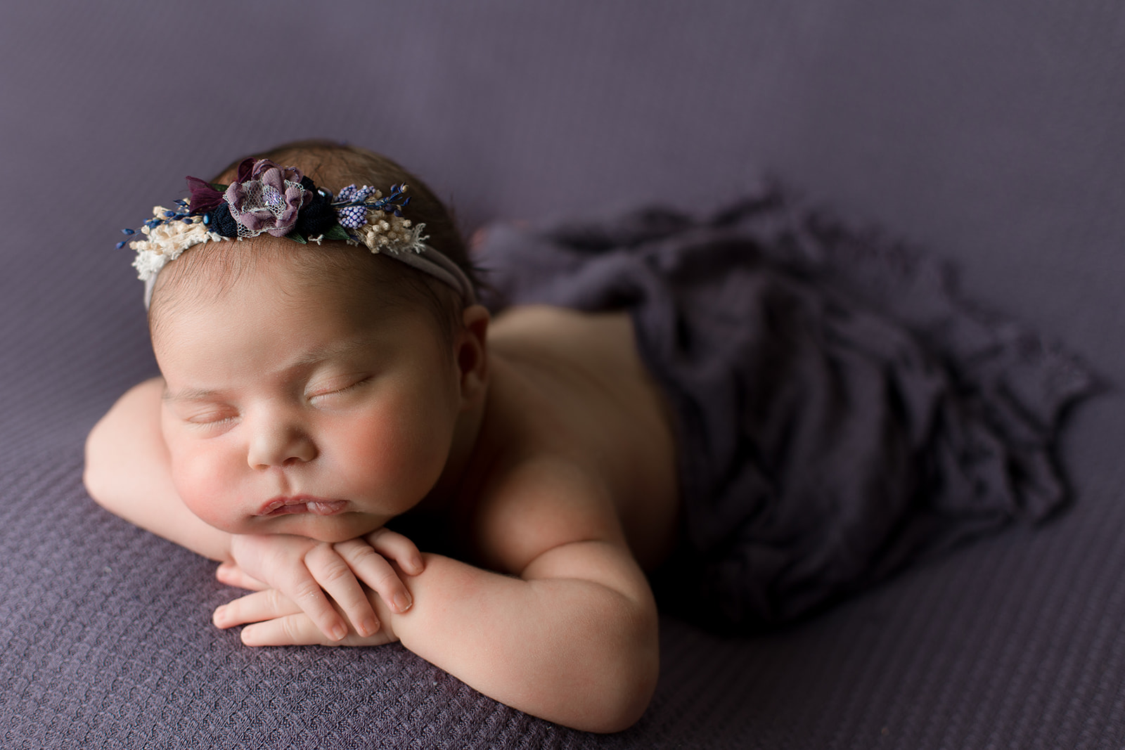 A newborn baby girl sleeps on her stomach on a purple bed in a matching floral headband after meeting Houston Babysitters