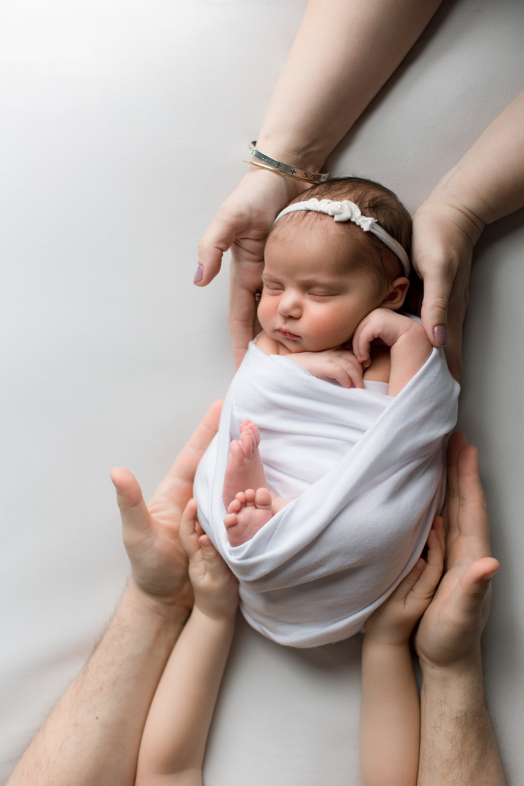 A newborn baby girl in a white swaddle sleeps in mom, dad's, and big sibling's hands