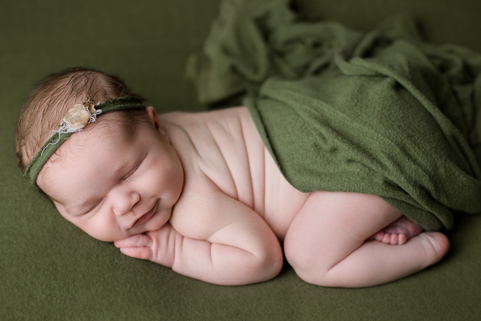 A newborn baby girl smiles in her sleep while covered with a loose green blanket before visiting Houston Preschools