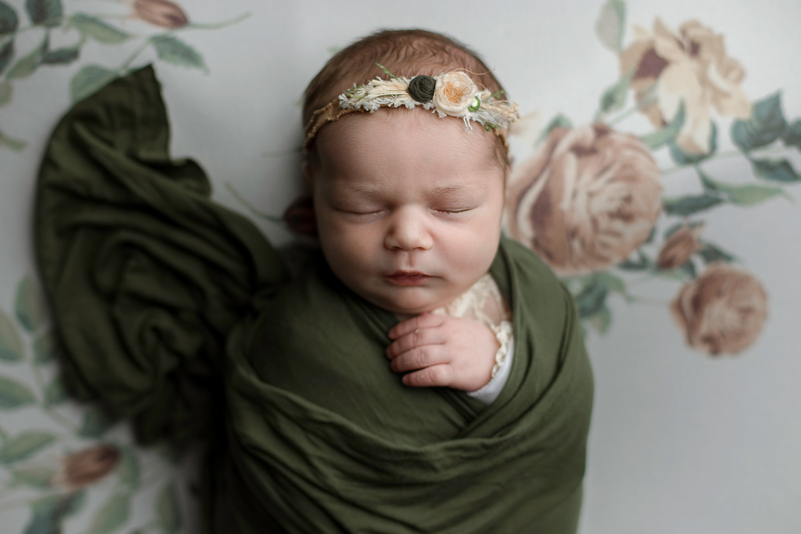 A newborn baby sleeps in a green swaddle on a floral bed with a floral headband after visiting Houston Preschools