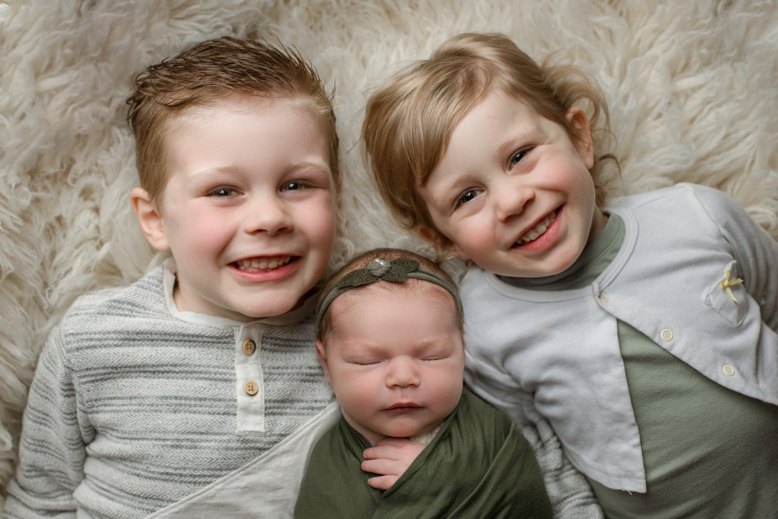 Happy toddler brother and sister lay on a fur rug with their sleeping newborn sister between them