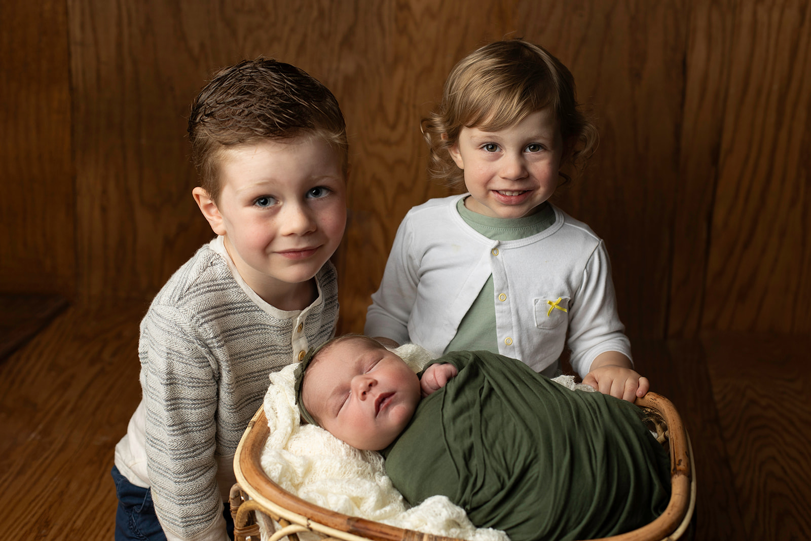 Toddler brother and sister smile over their sleeping newborn baby sister in a wicker basket after some Indoor Activities For Toddlers Houston